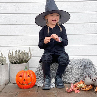 A young girl dressed as a witch sits on a bench wearing a smartwatch, smiling with a pumpkin beside her. The smartwatch ensures safe trick-or-treating with GPS tracking and communication.