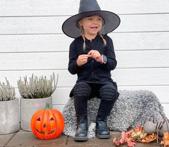 A young girl dressed as a witch sits on a bench wearing a smartwatch, smiling with a pumpkin beside her. The smartwatch ensures safe trick-or-treating with GPS tracking and communication.