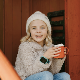 Smiling child with blonde hair wearing a white beanie and fuzzy jacket, holding a red mug with both hands. The child is wearing an Xplora smartwatch on their wrist while sitting in a cozy outdoor setting.