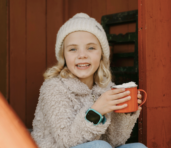 Smiling child with blonde hair wearing a white beanie and fuzzy jacket, holding a red mug with both hands. The child is wearing an Xplora smartwatch on their wrist while sitting in a cozy outdoor setting.