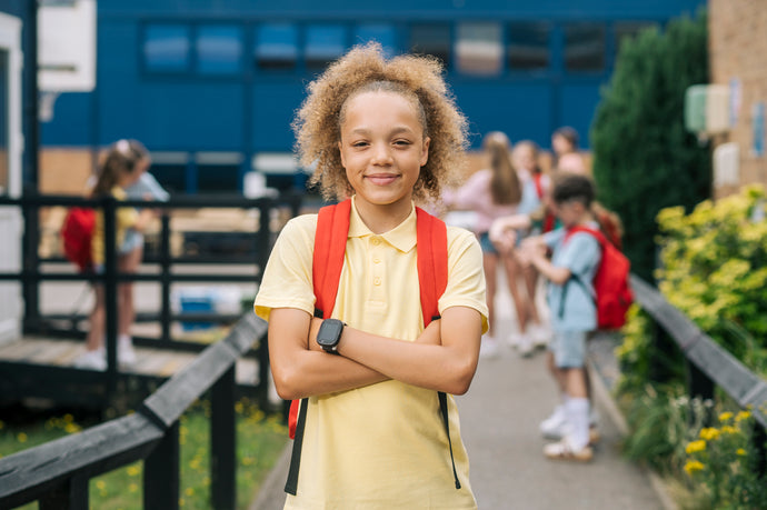 Young girl with a red backpack standing in front of a school with other students in the background.
